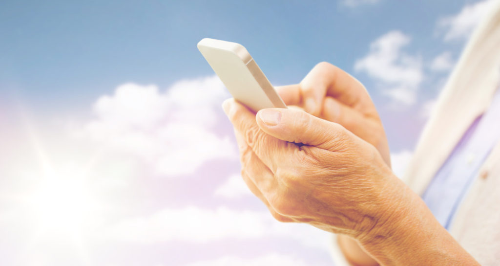 technology, age and people concept - close up of senior woman hands with smartphone texting message over blue sky and clouds background