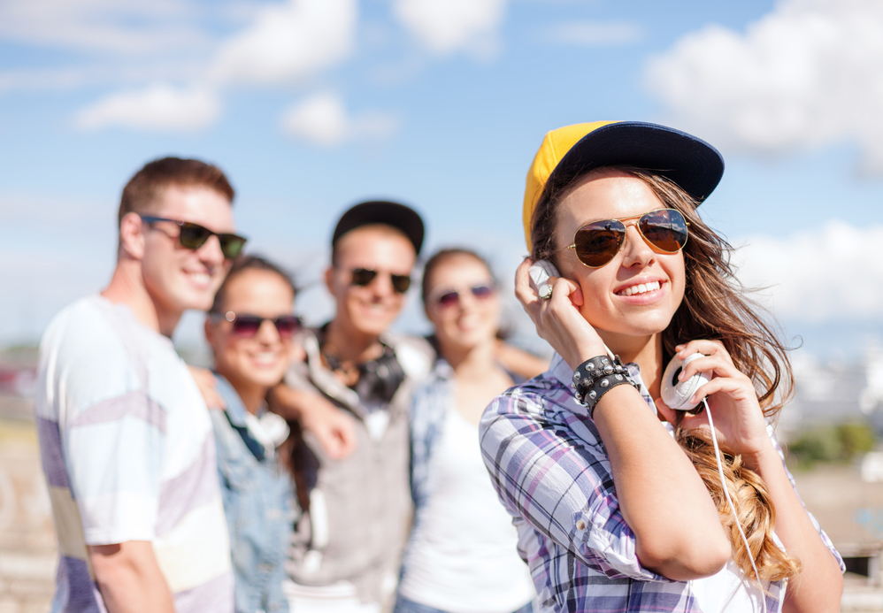 summer holidays and teenage concept - teenage girl in sunglasses, cap and headphones hanging out with friends outside